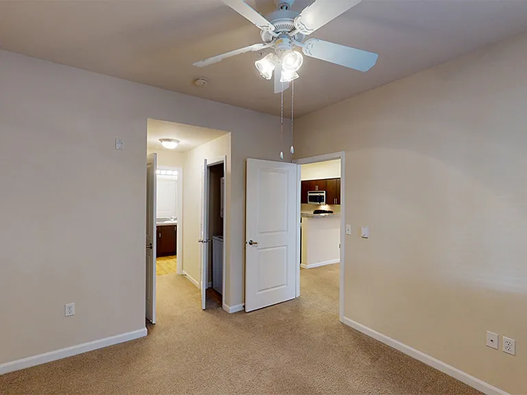 Carpeted apartment bedroom looking into kitchen and bathroom areas.