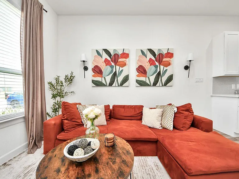 Comfortable living area with a sectional sofa, coffee table, and large window with blinds and curtains, adjacent to the kitch