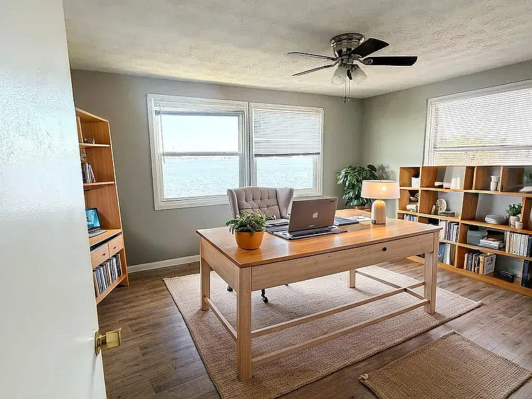 Bright and inviting home office with elegant wooden desk, chic gray chair, and ample shelving space, offering serene water views through large windows.