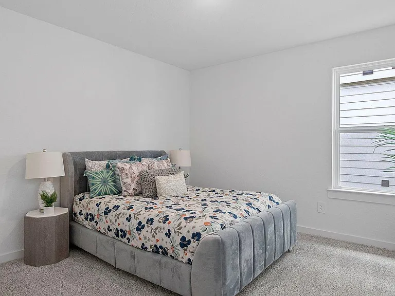 Staged bedroom with an upholstered bed, bedside tables and lamps, plush carpet, and a large window for natural light.
