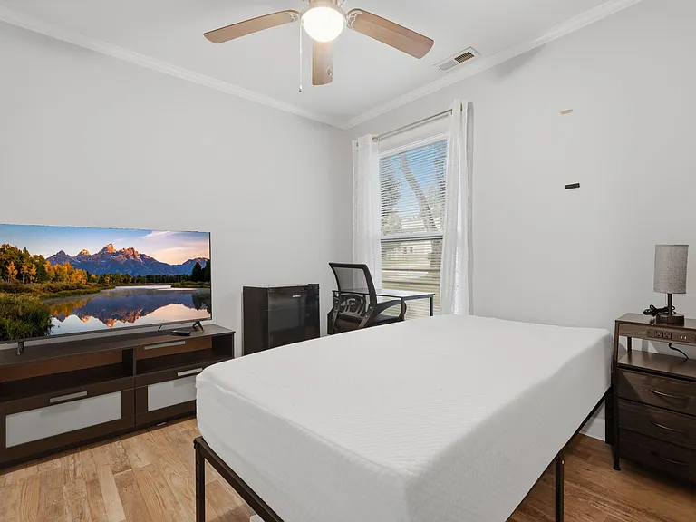 Furnished bedroom with a full bed, nightstand, TV stand, and a compact desk area set by a window with blinds.