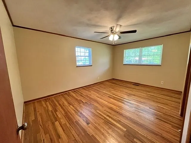 Large bedroom with beautiful hardwood flooring, neutral paint colors, and abundant natural light from multiple windows. This spacious room offers a bright and airy feel with plenty of space for a full bedroom setup. Features include warm wood trim, a mode