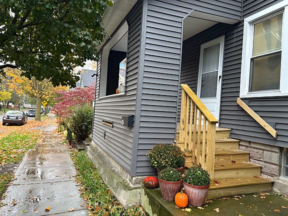The front entrance to 45 Luzerne, showing porch and steps, vinyl siding, door and screen door, windows, and mailbox (all new 2025). A row of perennial flowers has since bloomed between the sidewalk and the structure. The potted plants are provided.