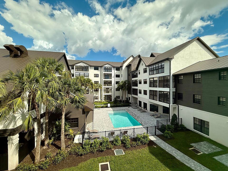 Vibrant apartment complex with a sparkling pool, surrounded by lush palm trees and modern architecture, basking under a bright blue sky.