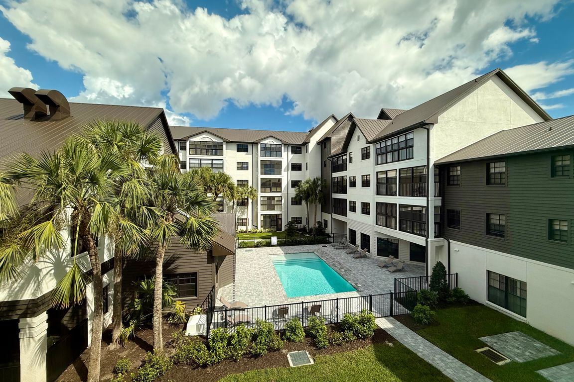 Vibrant apartment complex with a sparkling pool, surrounded by lush palm trees and modern architecture, basking under a bright blue sky.