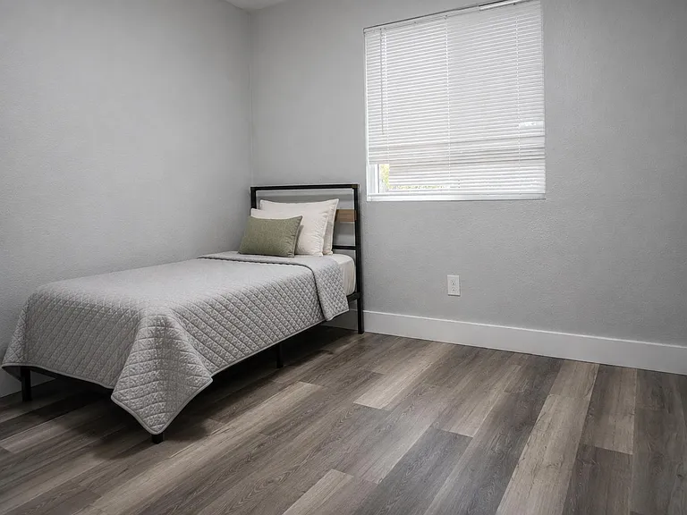 Private bedroom with a twin bed, wide-plank flooring, and a window with blinds for comfortable natural light and privacy.