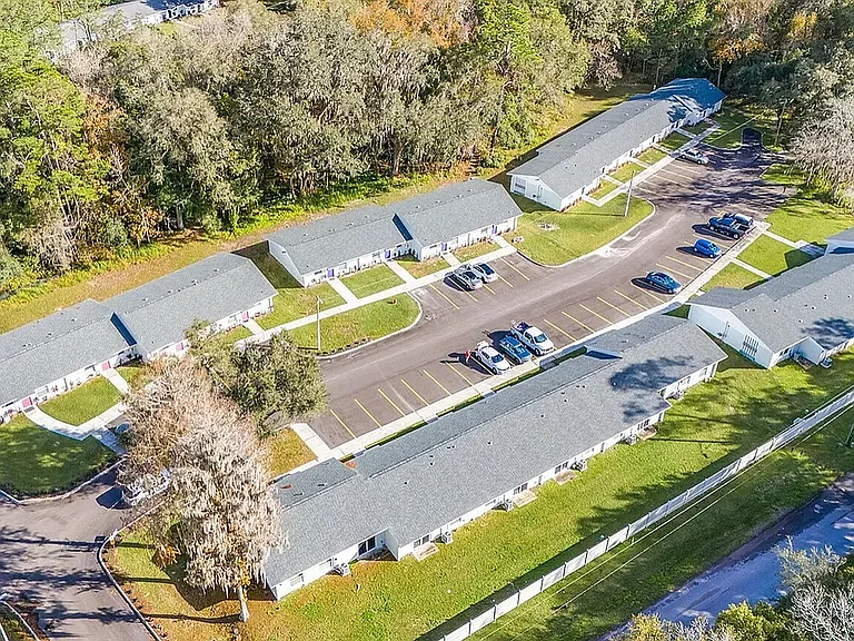 Charming aerial view of a well-maintained apartment complex with spacious parking and lush green surroundings, nestled beside a serene wooded area.