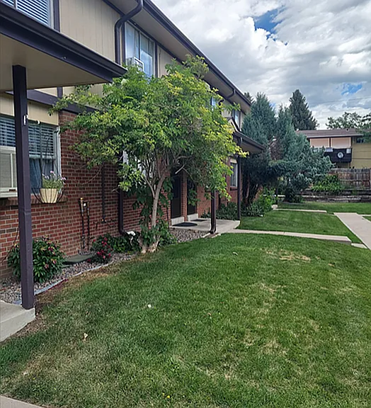 Charming apartment exterior with lush green lawn, vibrant flowers, and inviting walkway under a bright, cloudy sky.