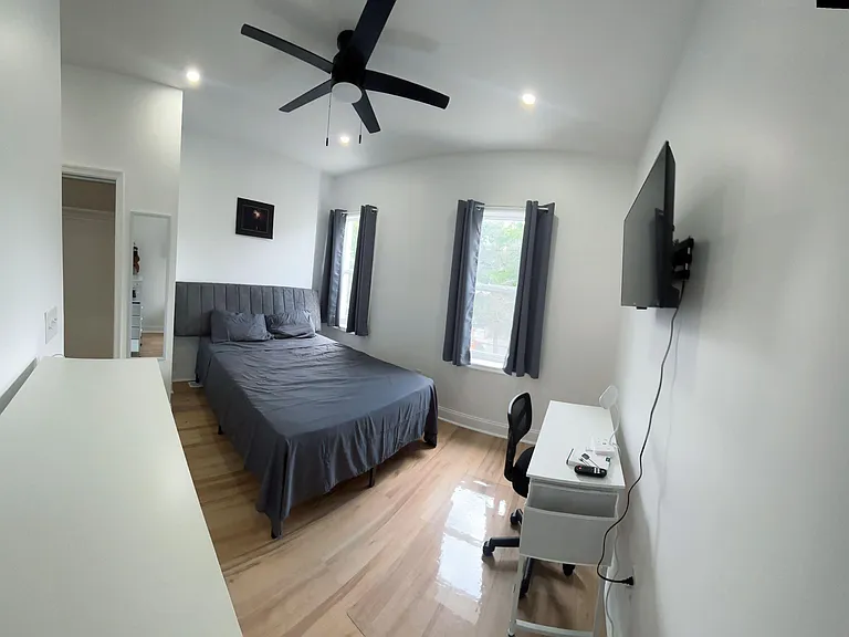 Private bedroom with vaulted ceiling, ceiling fan, two windows, and a work desk; includes wall-mounted TV and closet access.
