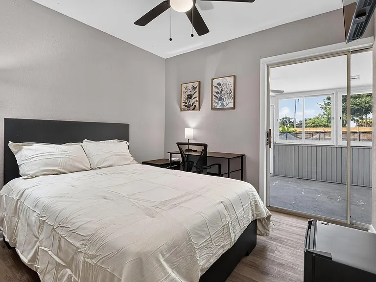 Furnished bedroom with ceiling fan, dedicated desk/work area, and sliding-door access to an enclosed porch.