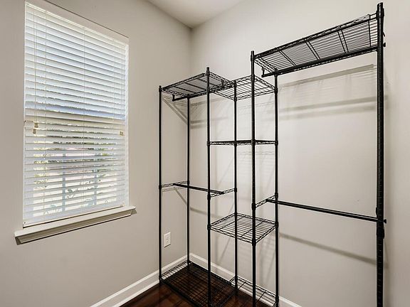 Compact room with a window and built-in blinds, plus freestanding shelving and hanging storage for clothes and gear.