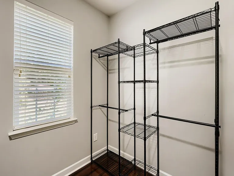 Compact room with a window and built-in blinds, plus freestanding shelving and hanging storage for clothes and gear.