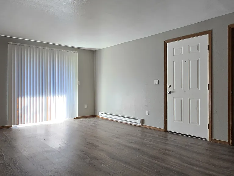 Bright, spacious living room with wood-style flooring, neutral tones, and large sliding glass doors for natural light. Includes a convenient entryway and extra storage closet, offering a clean, modern space ready for your personal touch.
