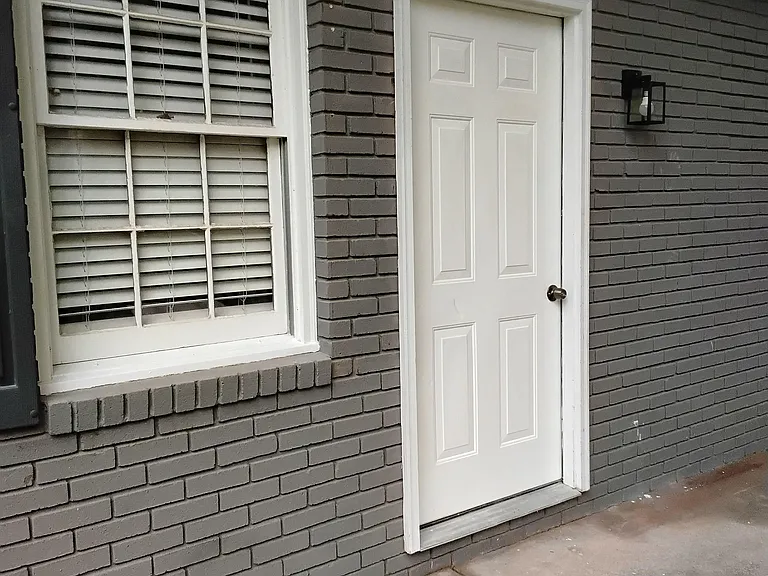 Covered exterior entry with a concrete walkway, brick facade, and secure door access next to a shaded window.