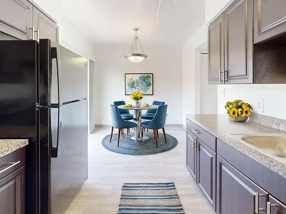 Bright and modern kitchen with sleek countertops, dark cabinetry, stainless steel sink, and adjacent dining area with elegant blue chairs and a stylish round table.