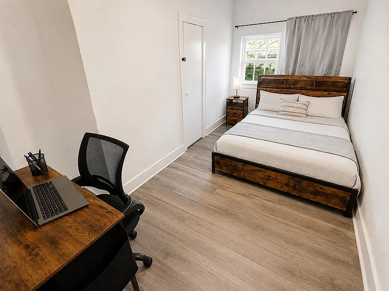 Furnished bedroom with a full bed, nightstand, and dedicated desk workspace, plus a window and closet for storage.