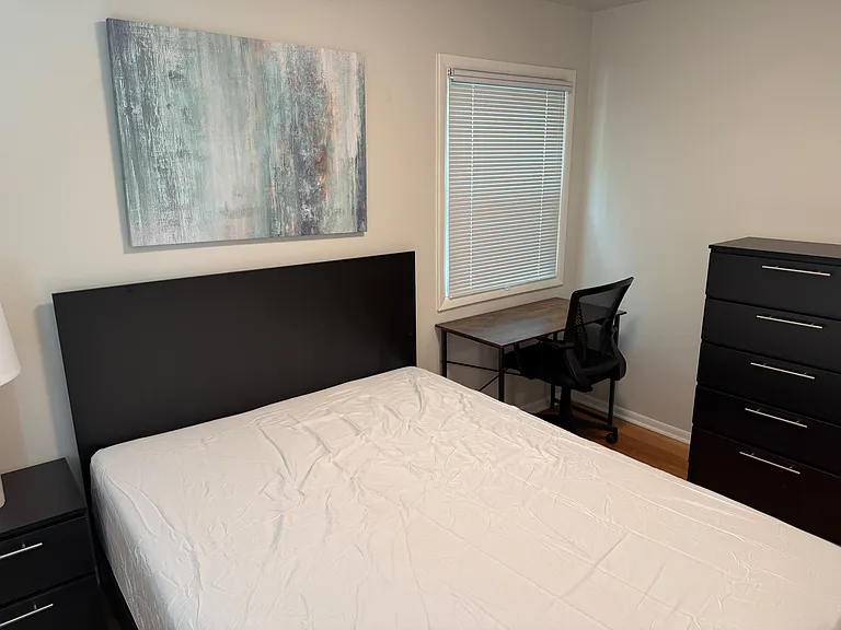 Furnished bedroom with a bed, dresser, and a dedicated desk by the window for working or studying.