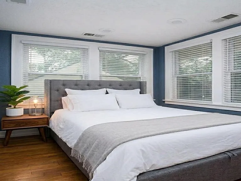 Spacious bedroom with a queen bed, hardwood-style floors, and large corner windows with blinds for natural light.