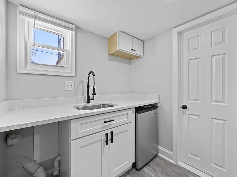Compact kitchenette with stone-look counters, undermount sink, shaker cabinetry, and a mini fridge with window light.