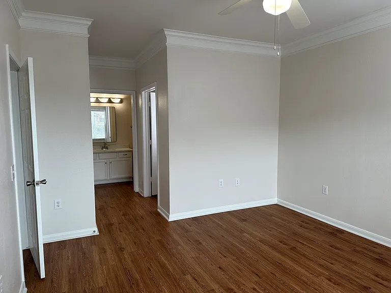 Bright and inviting empty room with elegant crown molding and wood-style flooring, leading to a cozy bathroom with ample natural light. Bright and inviting empty room with elegant crown molding and wood-style flooring, leading to a cozy bathroom with ampl