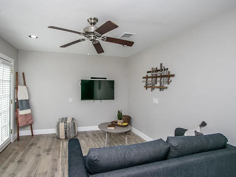 Detached casita living area at, Tempe rental home. Features a wall-mounted flat-screen TV, a dark grey sofa, a round table, patterned geometric pouf, and a decorative blanket ladder near the glass doors. Comfortable media space for a top Tempe, AZ stay.