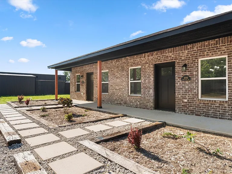 Brick exterior featuring a covered porch with wood posts, concrete walkway, and landscaped garden beds with stepping stones