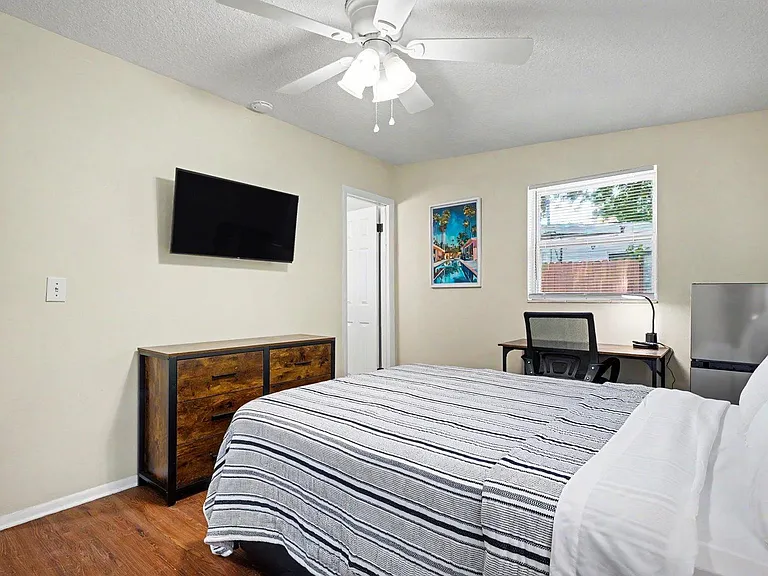 Large bedroom with a ceiling fan, wall-mounted 40" TV, 6-drawer dresser, work desk w/lamp, and mini fridge.