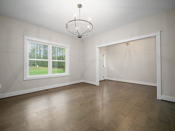 An empty room with light gray walls, hardwood floors, large window, and modern light fixtures. Doorw
