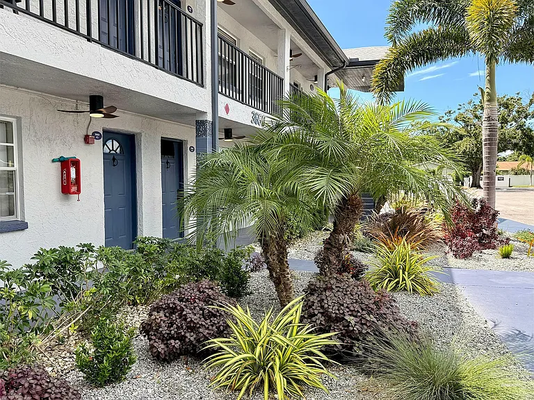 Charming apartment entrance with lush landscaping, featuring vibrant tropical plants under a clear blue sky.