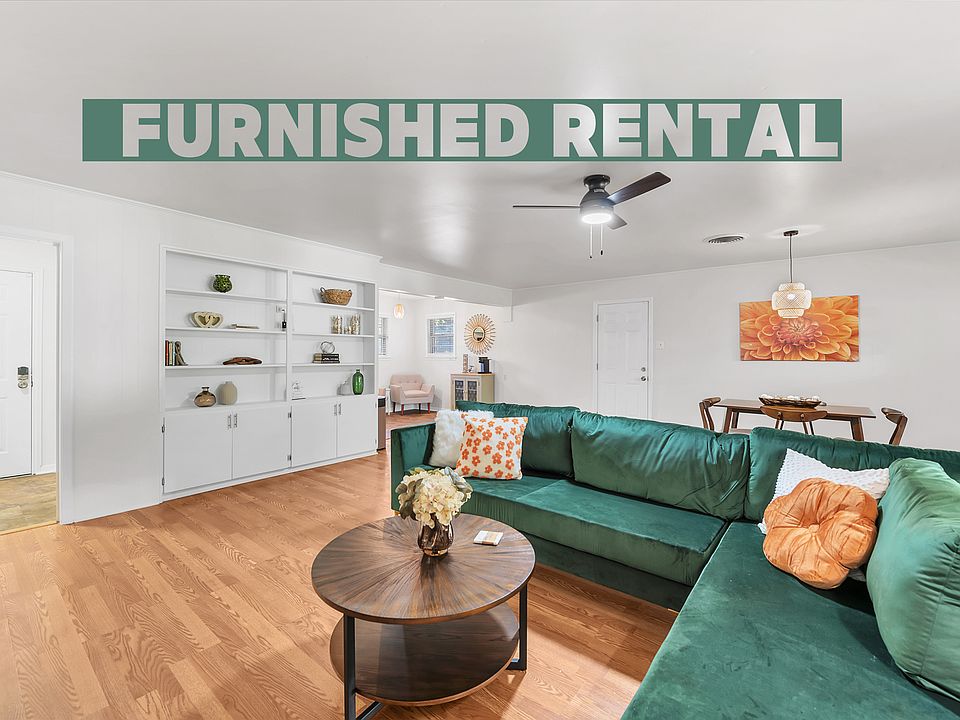 A vibrant living room featuring an emerald-green sectional, warm wood flooring, orange curtains framing a sliding glass door, and a sleek ceiling fan. A wooden media console, HD TV, and colorful artwork complete the cozy, stylish, and inviting space.