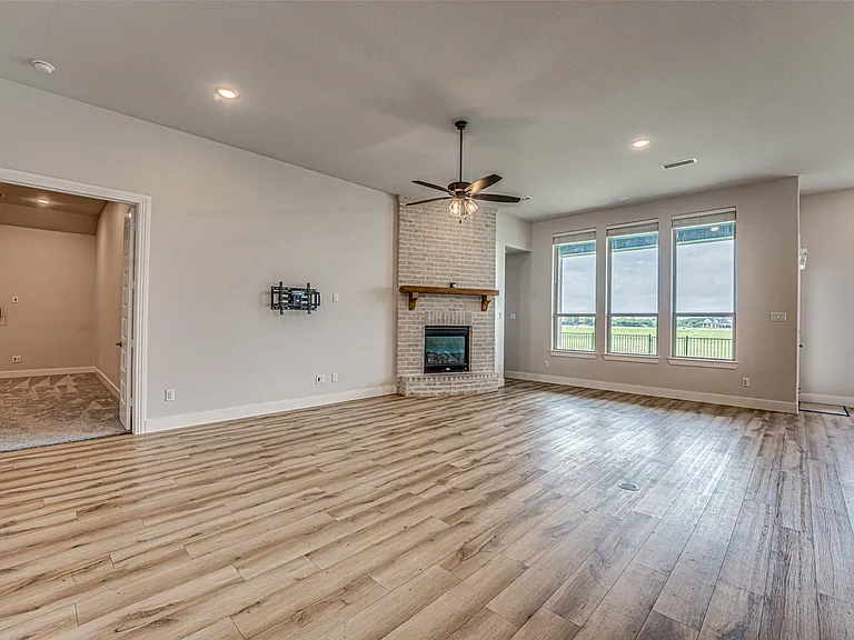 Spacious living area featuring wood-finish flooring, a white brick fireplace with a wood mantel, a ceiling fan, and multiple windows with roller shades
