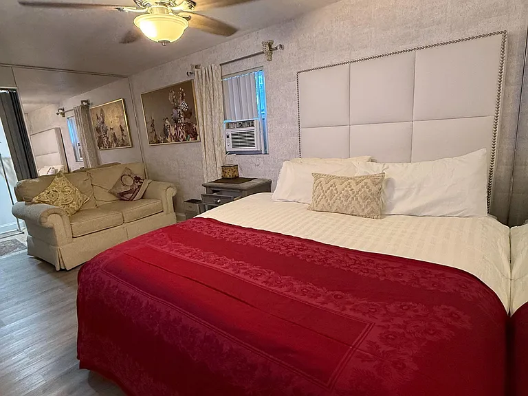 Spacious furnished bedroom with a large bed, seating area, ceiling fan, and window A/C for added comfort.