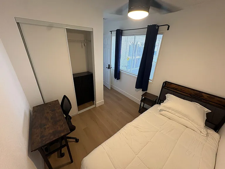 Furnished bedroom with a bed, nightstand, and desk, plus a sliding-door closet and window with blinds and curtains.