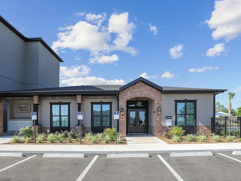 Charming clubhouse exterior with stylish brick facade, manicured landscaping, and ample parking under a clear blue sky.