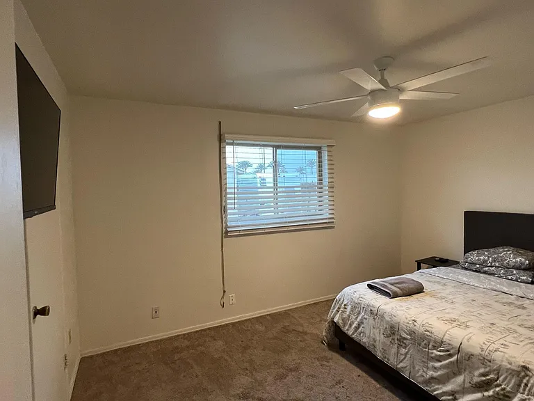 Spacious bedroom with carpeted floors, ceiling fan, large window with blinds, and a wall-mounted TV for easy viewing.