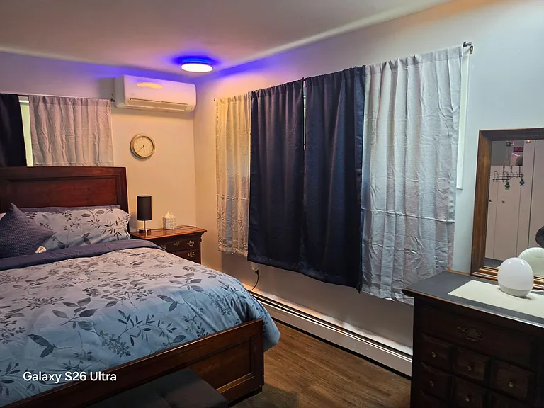 Furnished bedroom with a full bed, nightstand, dresser, and window coverings, plus a wall-mounted mini-split for comfort.