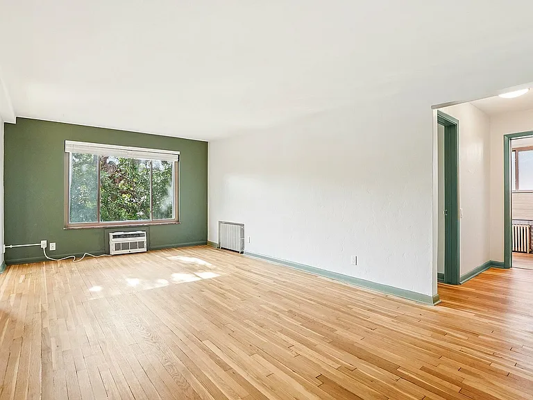 Bright and airy living room with hardwood floors, large window offering lush views, and fresh white and green walls.