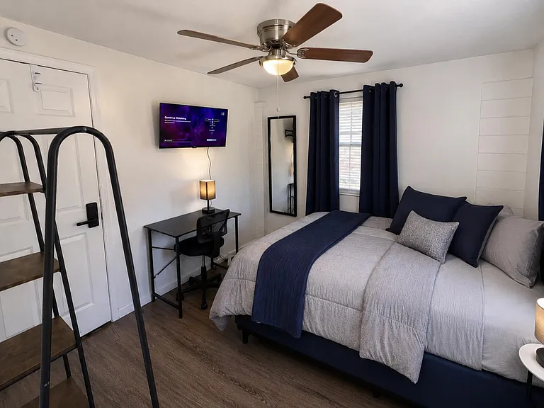 Furnished bedroom with a full bed, desk workspace, ceiling fan, and wall-mounted TV beside a window with curtains.