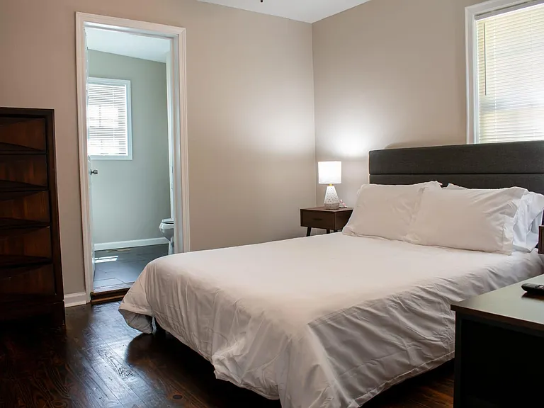 Spacious bedroom with a comfortable bed, nightstands, dresser storage, and an en-suite bath plus window blinds.