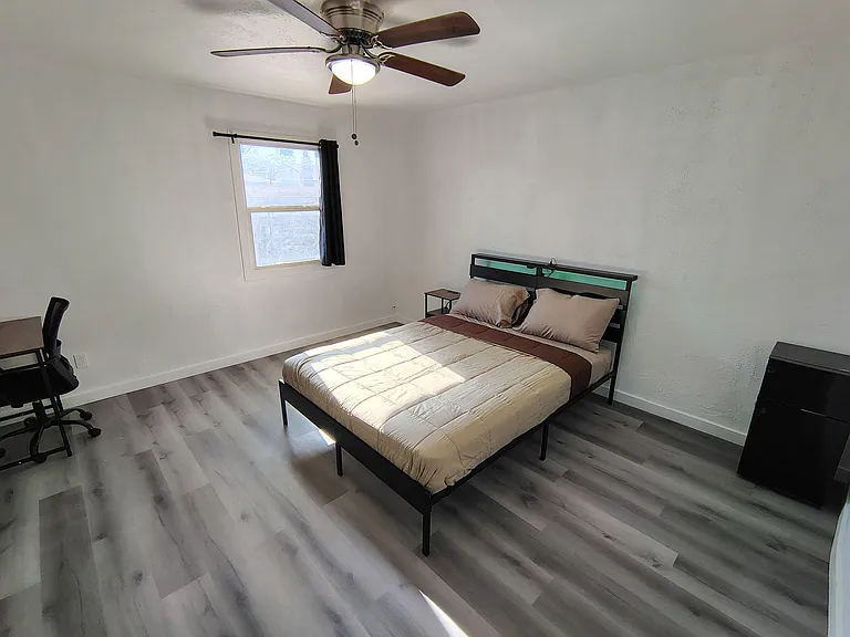 Furnished bedroom with a full bed, nightstand, and dedicated desk space, plus a ceiling fan and window for natural light.