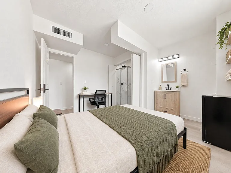 Spacious furnished bedroom with a work desk, mini fridge, and an en-suite bath featuring a walk-in shower and vanity storage.