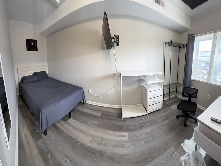 Furnished bedroom featuring a bed, wall-mounted TV, desk with chair, and open shelving for storage near a window.