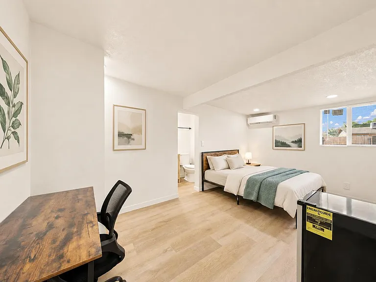 Furnished bedroom with desk workspace, mini fridge, and wall-mounted A/C, plus an ensuite bath and window with blinds.