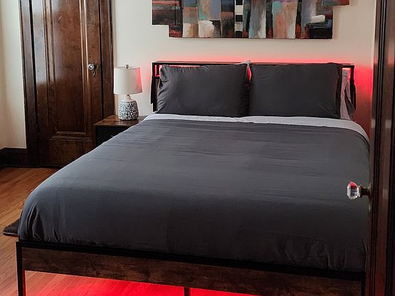 Bedroom with queen sized bed that includes LED lights and a charging station, desk, chair, file cabinet, shelf unit and large closet with mirror.