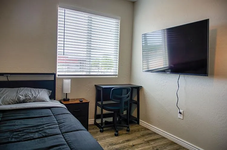 Private bedroom with a full bed, nightstand, and a dedicated desk area, plus a wall-mounted TV and window blinds.