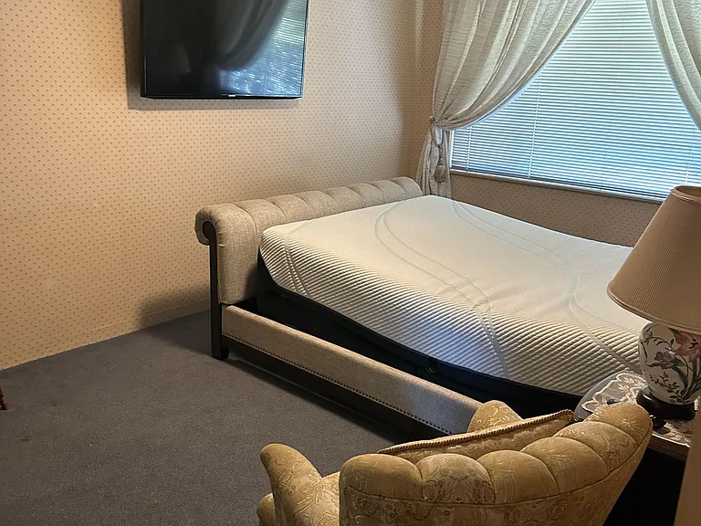 Furnished bedroom with a cushioned bed frame, wall-mounted TV, ceiling fan, and a window with blinds and curtains.