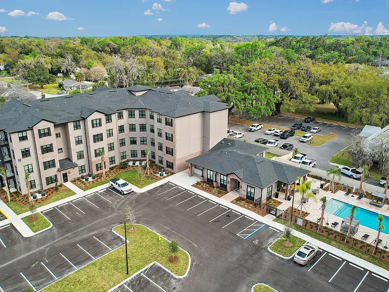 Aerial view of a modern apartment complex with a lush green backdrop, featuring a welcoming courtyard, ample parking, and a sparkling pool area.