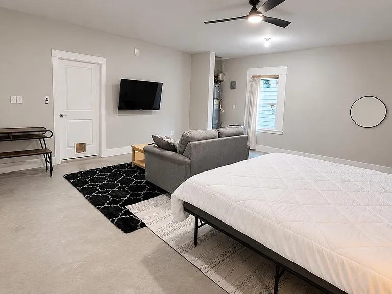 Spacious studio-style bedroom with ceiling fan, wall-mounted TV, seating area, and a window for natural light.