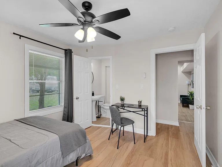 Furnished bedroom with a ceiling fan, large window with blinds, and a dedicated desk area, plus easy access to an ensuite bat