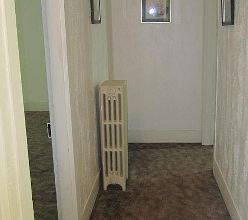 Hallway at top of stairs. Doorway to living room on left. Small bedroom at end of hallway to the right. Kitchen is to the immediate right, at the top of the stairs.